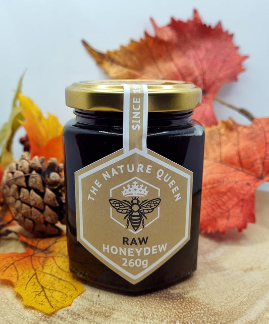 A jar of raw honeydew honey labeled 'The Nature Queen Raw Honeydew 260g' with autumn leaves and a pine cone in the background.